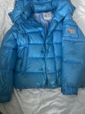 Blå pufferjacka från Moncler - Säljer en riktigt fet ljusblå pufferjacka från Moncler med glansig finish och klassisk logga på ärmen. Jackan har stora, vadderade paneler, dragkedja och tryckknappar samt fickor med dragkedja. Perfekt för kalla vinterdagar och riktigt snygg streetstil.