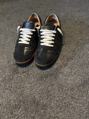Gats - Snygga svarta sneakers med vita skosnören och brun sula. Ovandelen är en mix av mocka och slätt läder för en stilren look. Låg modell som passar perfekt till jeans eller chinos. Insidan är fodrad i beige läder för extra komfort.
