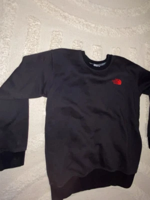 Svart sweatshirt från The North Face - Säljer en svart sweatshirt från The North Face med röd broderad logga på bröstet. Tröjan har rund halsringning, långa ärmar och ribbade muddar vid ärmslut och nederkant. Perfekt för en avslappnad och stilren look.