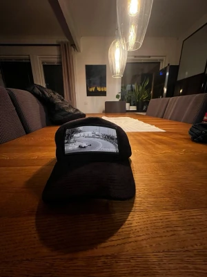 Svart keps från Vernando med motiv - Säljer en svart keps från Vernando med ett coolt svartvitt fotomotiv framtill som visar en racingbana. Kepsen har klassisk böjd skärm och mesh baktill för extra ventilation. Justerbar snapback för perfekt passform. Perfekt accessoar för en sportig och avslappnad stil.