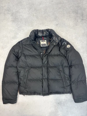 Svart dunjacka från Moncler - Snygg svart dunjacka från Moncler med hög krage och två dragkedjefickor framtill. Jackan har ett quiltat mönster och Moncler-logga på ärmen. Materialet är glansigt på vissa partier vilket ger en cool kontrast. Perfekt för kalla vinterdagar.