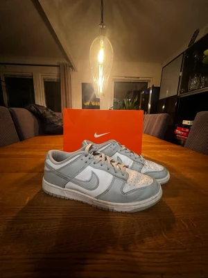 Nike Dunk Low vit och ljusgrå - Nike Dunk Low sneakers i vitt och ljusgrått läder med klassisk Swoosh på sidan. Skorna har vit tåbox med perforeringar, ljusgrå snörning och yttersula. Perfekt för dig som gillar stilrena och tidlösa sneakers från Nike.