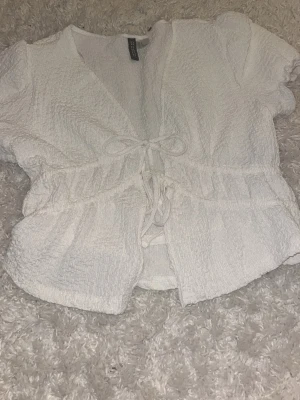 Vit croppad topp med struktur - Säljer en vit croppad topp med korta ärmar och snygg struktur i tyget. Toppen har en lätt rynkad effekt och markerad midja som ger en trendig siluett. Perfekt att ha en sommarkväll🤍har bara testa den på en gång!💞