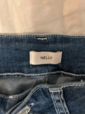 Blå jeansshorts från Nelly - Säljer ett par blå jeansshorts från Nelly med rå kant och dekorativa sömmar i vitt på bakfickorna. Klassisk femficksmodell med knappstängning och bälteshällor. Perfekta för varma dagar och enkel att matcha med olika toppar.
