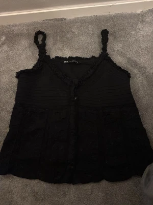 Svart spetslinnetopp från Zara - Säljer en svart linnetopp från Zara med spetsdetaljer och knappar framtill. Toppen har smala axelband och en lätt volangkant vid urringningen. Perfekt för dig som gillar en feminin och trendig stil.