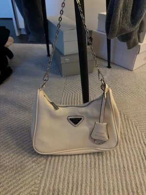 Beige väska silver/svart detaljer - Vit axelväska i kompakt modell med kedjerem i metall. Mjuk yta, dragkedjestängning och detaljer i silverfärgad metall. Smidig storlek som rymmer det viktigaste och sitter snyggt både på axeln och crossbody med längre kedja.