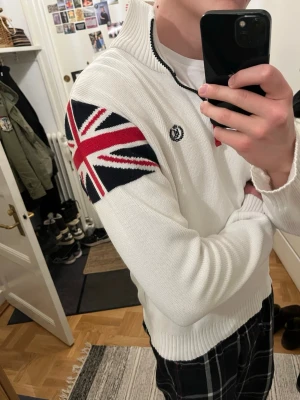 Henri Lloyd Union Jack halfzip  - Vit stickad tröja från Henri Lloyd med halv dragkedja och krage. Union Jack-flagga i rött, blått och vitt på ena ärmen och HL broderad på bröstet. Ribbstickade muddar och nederkant ger en snygg passform. Perfekt för dig som gillar preppy stil.