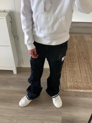 Svarta jeans med färgstänk - Säljer ett par svarta jeans med vita färgstänk och avslappnad passform. Jeansen har raka ben och sitter snyggt på höften. Perfekta för dig som vill ha en streetwear-inspirerad look. Använda 2 gånger , fick i present 