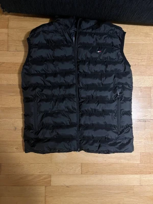 Svart dunväst från Tommy Hilfiger - Snygg svart dunväst från Tommy Hilfiger med diskret logga på bröstet. Västen har horisontella sömmar, dragkedja framtill och två fickor med dragkedja. Perfekt för lager-på-lager och håller dig varm under kyliga dagar.