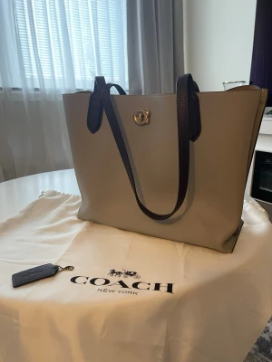 Coach väska - Stilren beige väska från Coach med mörkbruna handtag och guldigt spänne. Väskan har ett rymligt huvudfack och en innerficka med dragkedja i klassiskt Coach-mönster. Tillverkad i slätt läder och perfekt för dig som gillar snygg och praktisk design. Jätte bra skick, som ny. Inga defekter. Bredd: 35cm, höjd: 28 cm, djup: 15 cm. 