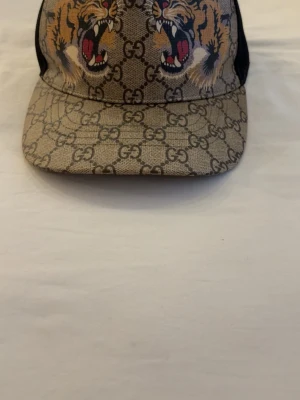 Gucci keps med tigertryck och GG-mönster - Säljer en beige Gucci keps med klassiskt GG-mönster och två stora tigerhuvuden framtill. Kepsen har svart mesh baktill och justerbart band med gröna och röda ränder. Materialet är canvas framtill och mesh bak. Perfekt för dig som vill sticka ut med en lyxig accessoar. Pris kan diskuteras 