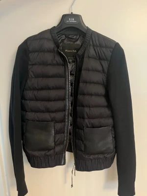 Svart pufferjacka från Massimo Dutti - Snygg svart cardigang från Massimo Dutti med quiltad front, släta ärmar och två stora fickor i skinnimitation. Jackan har dragkedja och rundad krage. Även en innerficka. Materialmix med både vadderad och stickad känsla. Kommer inte till användning. Storlek xs men passar också s.