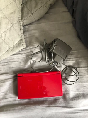 Röd Nintendo DS Lite med laddare - Säljer en röd Nintendo DS Lite med tillhörande laddare. Konsolen har dubbla skärmar, klassiska knappar och stilren vikbar design. Perfekt för dig som vill spela DS-spel på språng eller hemma. Färgen är riktigt snygg och sticker ut bland andra handhållna konsoler. OBS!! Ingen penna och e osäker på mic.