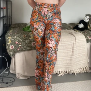 Färgglada blommiga leggings - Säljer ett par höga, utsvängda leggings med färgglatt blommönster i orange, rosa, blått och grönt. De har bred resår i midjan och är sydda i ett stretchigt, ribbat material som sitter skönt och följsamt. Perfekt för den som gillar lite 70-tals vibbar. Otroligt stretchiga!! Storlek XS men passar även mig som generellt har L i byxor, om än lite tighta i midjan