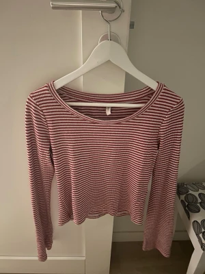 Randig långärmad topp i rött och vitt - Säljer en långärmad topp med smala röda och vita ränder. Toppen har en rund halsringning och är croppad i modellen. Perfekt till jeans eller kjol för en avslappnad look.
