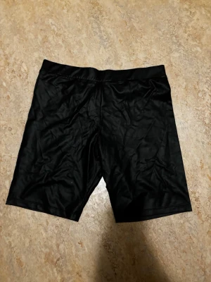 Svarta cykelshorts från H&M - Säljer ett par svarta cykelshorts från H&M. De har en enkel och clean design med elastisk midja och är tillverkade i ett stretchigt syntetmaterial som sitter skönt på kroppen. Perfekta till sommaren eller träning!