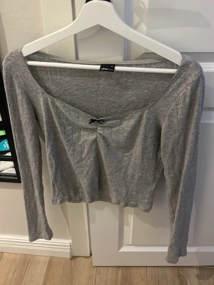 Grå ribbad topp med rosett - Snygg grå ribbad topp från gina tricot med lång ärm och liten svart rosett vid ringningen. Toppen har en croppad passform och är tillverkad i mjukt bomullsmaterial. Perfekt för en trendig och avslappnad look.