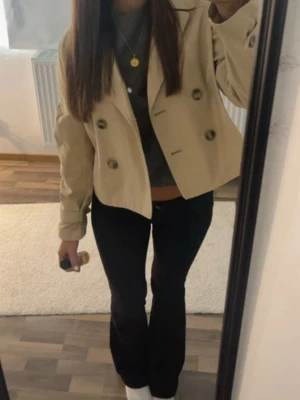 Beige kort trench coat  - Stilren beige kort trench coat med dubbelknäppning och stora knappar framtill. Klassisk krage och rak passform, perfekt för lager-på-lager. Tillverkad i ett lätt tyg som passar bra till vår och höst.