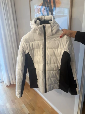 Vit och svart pufferjacka med huva - Snygg skidjacka i vitt med svarta paneler längs sidorna och ärmarna. Jackan har en hög krage, fast huva och dragkedja framtill. Två sidofickor med dragkedja och quiltad design ger en modern look. Perfekt för kalla vinterdagar.