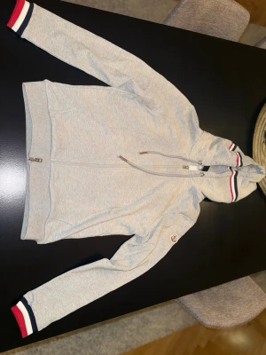Grå Moncler hoodie med ränder - ( VID KÖP FINNS DE BILD PÅ KVITTO FRÅN NK) ( NY PRIS MELLAN 4-5k)Snygg grå hoodie från Moncler med dragkedja framtill och huva med dragsko. Ärmsluten och huvan har stilrena ränder i rött, vitt och blått. Klassisk Moncler-logga på ärmen och två fickor fram. Perfekt för en chill och sportig look.