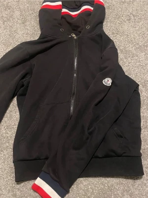 Svart Moncler hoodie med dragkedja - Snygg svart hoodie från Moncler med dragkedja framtill och klassisk Moncler-logga på ärmen. Huva med röda och vita ränder samt matchande muddar vid ärmslut. Tillverkad i mjukt bomullsmaterial för en bekväm känsla. Perfekt för en avslappnad och trendig stil.