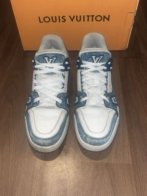 Louis Vuitton vita och blå sneakers - Snygga Louis Vuitton sneakers i vitt och blått med klassisk LV-logga på plösen och sidan. Skorna har denimliknande detaljer, vita snören och robust sula. Materialet är en mix av läder och textil, vilket ger en exklusiv och trendig look.