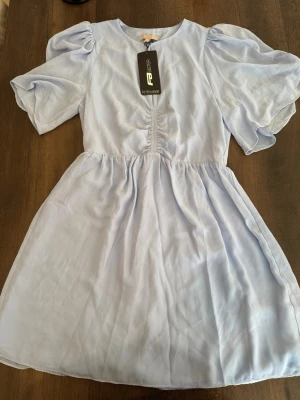 Ljusblå kortklänning med puffärm - Supersöt ljusblå kortklänning från Resteröds med puffiga, korta ärmar och lätt rynkad detalj framtill. Klänningen har en flowy kjol och är tillverkad i ett tunt, mjukt material som känns luftigt och somrigt.