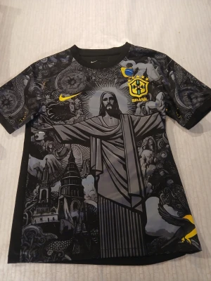 Nike Brasil t-shirt med tryck - Snygg svart t-shirt från Nike med Brasil-logga och stort grafiskt tryck av Kristusstatyn och detaljer i gult. T-shirten har korta ärmar och rund hals. Perfekt för dig som gillar unika och sportiga plagg med cool design.