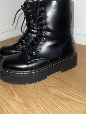 Svarta chunky kängor med snörning - Säljer ett par svarta kängor med högt skaft och grov platåsula. Kängorna har snörning hela vägen upp och är tillverkade i blankt konstläder. Perfekta för dig som gillar en edgy och trendig stil.