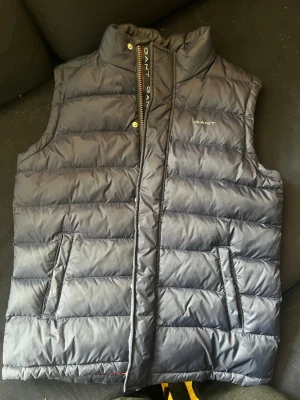 Mörkblå dunväst från Gant 158/164 - Snygg mörkblå dunväst från Gant med diskret logga på bröstet och cool röd insida. Västen har ståkrage, dragkedja och tryckknappar samt två praktiska fickor framtill. Perfekt för lager på lager och streetwear-vibe. Storlek 158/164