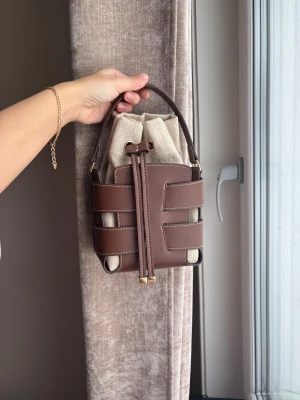 Brun bucket bag med canvasdetaljer - Trendig handväska i bucket-modell med brunt konstläder och beige canvasinsats. Väskan har ett unikt flätat lädermönster, kort handtag och dragskostängning. Perfekt accessoar för en modern look.