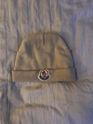 Beige mössa med logga mnclr  - Säljer en beige ribbstickad mössa med uppvikt kant och klassisk logga framtill. Mössan är enkel och stilren, perfekt för att hålla värmen under kalla dagar. Materialet känns mjukt och bekvämt mot huden.