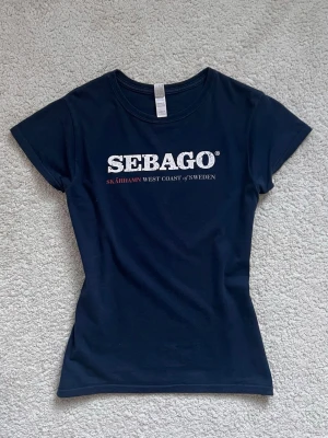 Sebago t-shirt - Snygg marinblå t-shirt från Sebago med vit och röd text 'SEBAGO SKÄRHAMN WEST COAST OF SWEDEN'. Står M men passar som S/XS
