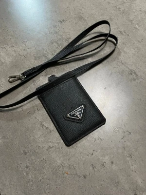 Svart korthållare med rem från HM - Snygg svart korthållare från Hm i läder med klassisk trekantig logga framtill. Kommer med en lång rem så du kan bära den runt halsen eller crossbody. Perfekt för kort och ID när du är på språng.