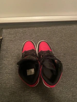 Svarta och röda Air Jordan 1 Mid - Säljer ett par Air Jordan 1 Mid sneakers i svart och rött med vita sulor. Klassisk design med svart swoosh, röda detaljer och svart snörning. Skorna har en högre siluett och är tillverkade i en kombination av läder och textil. Perfekta för dig som gillar streetwear och ikoniska sneakers.