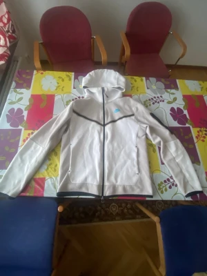 Ljusgrå Nike tech hoodie med dragkedja  - Snygg ljusgrå hoodie från Nike med hel dragkedja framtill och svart detaljsöm. Huva med dragsko och två sidofickor med dragkedja. Tillverkad i mjukt material, perfekt för en avslappnad stil. Liten blå logga på bröstet.