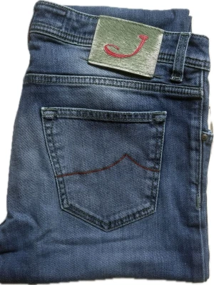 Jacob Cohën Jeans | W35 - Snygga blå jeans från Jacob Cohen med klassisk femficksmodell och röda detaljer på bakfickan och vid knäppningen. Jeansen har en grön patch med rött J på linningen och silverfärgade knappar. Tillverkade i mjukt denimtyg för en bekväm känsla.
