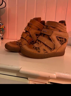 Bruna Carinii sneakers med leoparddetaljer - Säljer ett par bruna sneakers från Carinii med coola leopardmönstrade detaljer på sidorna. Skorna har tre kardborreband och en högre sula som ger extra attityd. Tillverkade i mocka och textil, perfekta för dig som vill sticka ut.