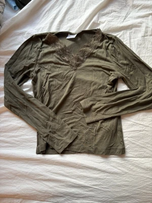 Olivgrön topp med spetsdetalj - Olivgrön långärmad topp med v-ringning och dekorativ spets längs halsringningen. Mjuk och stretchig känsla, perfekt för en avslappnad men ändå stilren look.