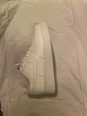Nike Air Force 1 vita sneakers - Klassiska vita Nike Air Force 1 sneakers i lågt utförande. Skorna har en stilren design med perforerad tå, vita skosnören och den ikoniska swoosh-loggan på sidan. Tillverkade i slätt läder med en rejäl gummisula för grym komfort och streetstyle.