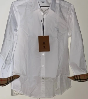 Burberry skjorta - Klassisk vit skjorta från Burberry med långa ärmar och knappar framtill. Ikoniska beige-rutiga detaljer syns på insidan av manschetterna. Skjortan har en liten broderad logga på bröstet och är tillverkad i bomull. Skjortan är oanvänd!