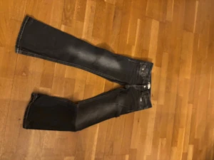 Mörkblå bootcut jeans med slitningar - Säljer ett par mörkblå bootcut jeans med snygga slitningar på låren och klassiska fem fickor. Jeansen har kontrastsömmar och detaljerade bakfickor med knappar. Perfekta för en avslappnad och trendig look.