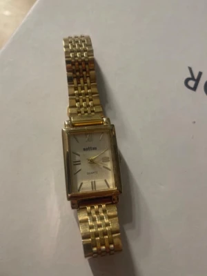 Guldfärgad kvadratisk klocka Soltas - Stilren guldfärgad klocka från Soltas med fyrkantig urtavla och klassiskt armband i metall. Urtavlan har romerska siffror och quartz-urverk. Perfekt accessoar för dig som gillar en elegant och tidlös look.