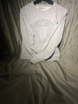 Grå sweatshirt med broderad tiger - Snygg grå sweatshirt från Tiger of sweden med det ikoniska broderade tigermotivet på bröstet. Tröjan har rund halsringning och är tillverkad i mjukt bomullsmaterial. Perfekt för en avslappnad och trendig stil.