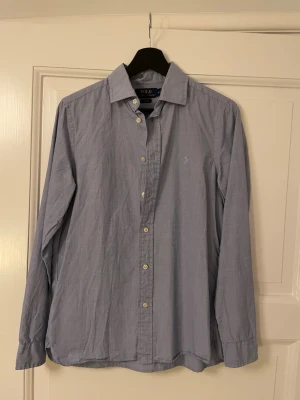 Blå skjorta från Polo Ralph Lauren - Klassisk blå skjorta från Polo Ralph Lauren med knappar framtill och lång ärm. Skjortan har en diskret broderad logga på bröstet och är tillverkad i bomull. Perfekt för en stilren och avslappnad look.