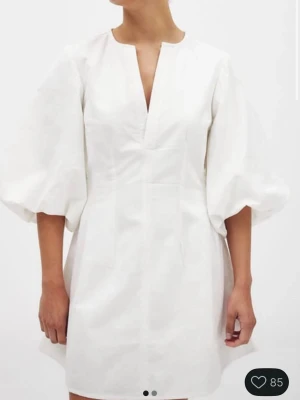 Studentklänning med puff ärmar - Stilren vit kort klänning med v-ringning och trekvarts puffärmar. Klänningen har en rak passform och är tillverkad i ett lätt bomullstyg som ger en fräsch känsla. Perfekt för dig som gillar minimalistisk och trendig stil.