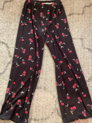 Svarta mjukis byxor med körsbärsmönster - Säljer ett par svarta loungewear-byxor med elastisk midja och ett gulligt körsbärsmönster i rött och grönt. Byxorna har en avslappnad passform och är perfekta för att chilla hemma. Materialet känns mjukt och svalt mot huden.