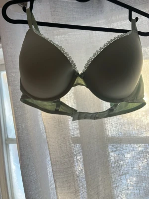 Ljusgrön bh - Snygg ljusgrön bh med vadderade kupor och dekorativ vit spetskant upptill. Bh:n har justerbara axelband och hakknäppning bak. Materialet är slätt och stretchigt för en bekväm passform. Den har 2 pytte pyttesmå fläckar på vänstra sidan men är ganska så säker på att de går bort med vanish 