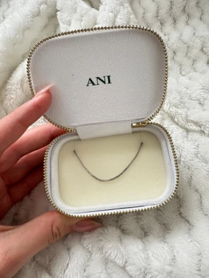 Silvrigt halsband från ANI - Snyggt och minimalistiskt halsband i silverfärgad metall från ANI. Kedjan är tunn och elegant med en liten grön detalj vid låset. Perfekt för dig som gillar stilrena accessoarer och vill ha något som funkar till många looks. Aldrig använd