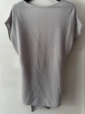 Grå topp - Snygg grå stickad topp med omlott-design på baksidan och korta ärmar. Toppen har en djup v-ringning baktil och är tillverkad i ett mjukt, ribbstickat material som ger en modern och avslappnad look.Anvönd en gång.